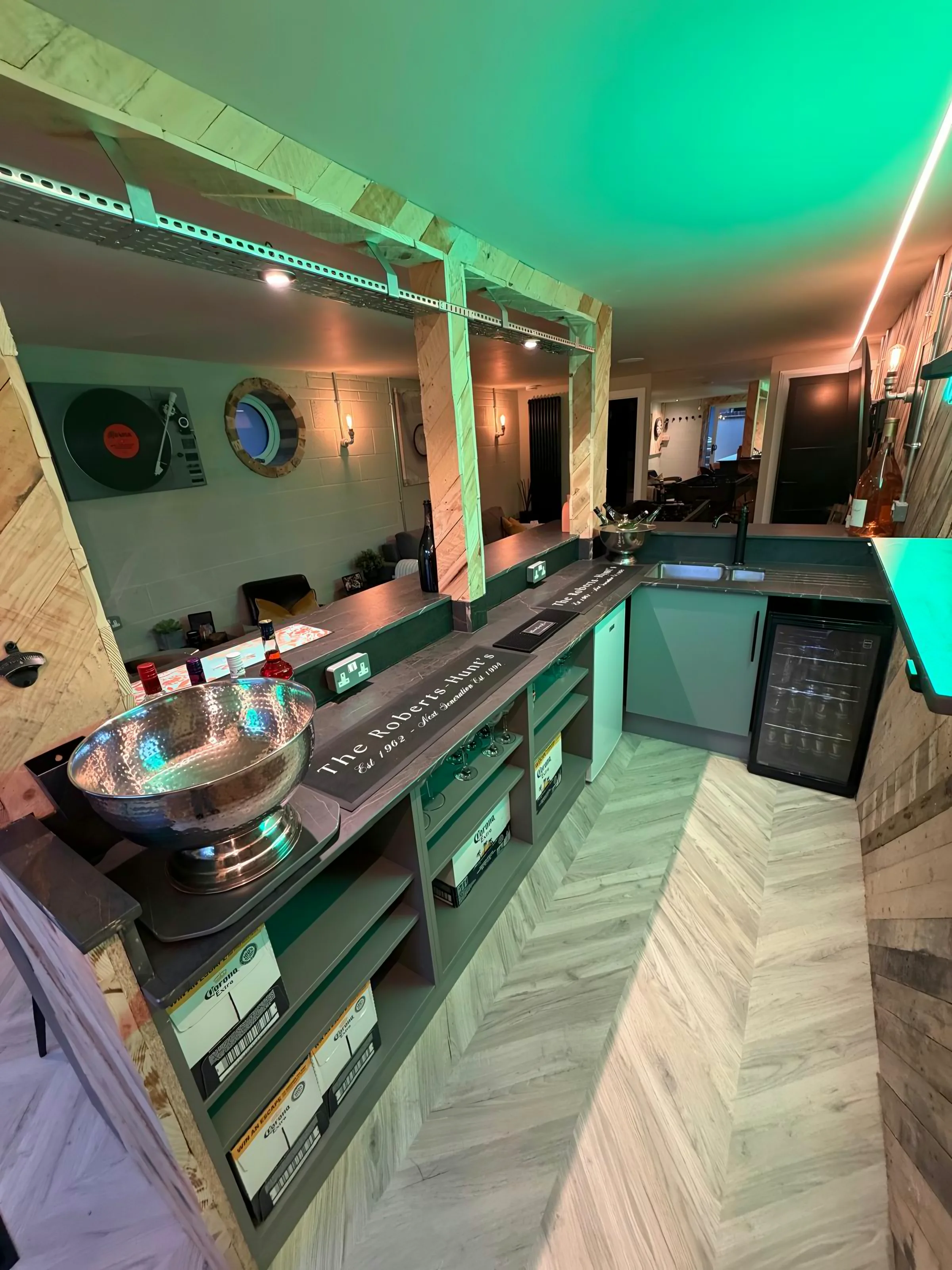 Black Laurent run down the length of a home bar under green cove lighting, bottles racked behind and a herringbone floor